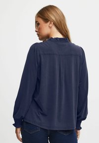 Navyblå blus med långa ärmar, volangkrage och plisserad ryggdetalj, tillverkad av mjukt tyg med åtsittande elastiska manschetter.