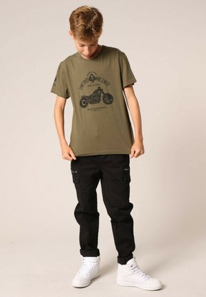 T-shirt olive avec un imprimé graphique noir d'une moto, associé à un pantalon cargo noir et des baskets blanches. L'enfant regarde vers le bas.