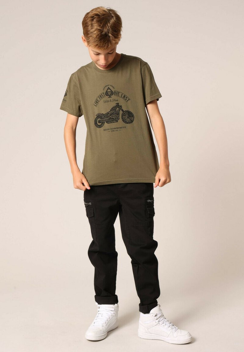 T-shirt olive avec un imprimé graphique noir d'une moto, associé à un pantalon cargo noir et des baskets blanches. L'enfant regarde vers le bas.