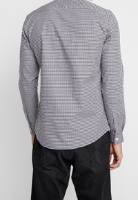 Uomo che indossa una camicia a maniche lunghe a scacchi piccoli bianco e nero e jeans neri, visto di spalle su uno sfondo neutro.