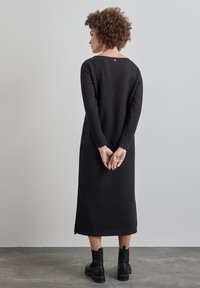 Robe noire à manches longues avec un large décolleté, en tissu texturé et fentes latérales. Associée à des bottines noires à la cheville. Petit détail de bouton à l'arrière.