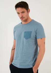 Světle modré bavlněné tričko s kulatým výstřihem, krátkými rukávy a texturovanou džínovou kapsou na levé straně hrudníku.