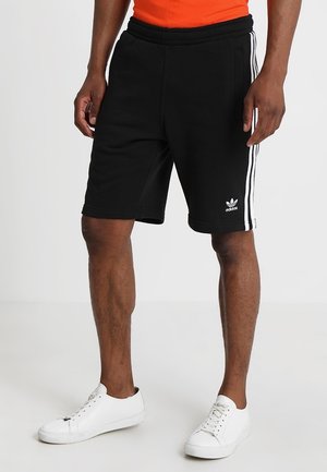 Mężczyzna ubrany w czarne spodenki Adidas z białymi bocznymi paskami i białe trampki, stojący na jednolitym jasnym tle.