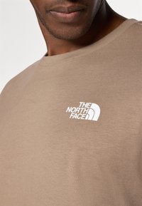 Tricou bej deschis din bumbac, cu un design cu gât rotund. Prezintă un logo alb în zona stângă a pieptului și o textură netedă.