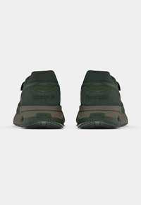 Um par de sapatilhas Reebok verde-escuras vistas de trás, mostrando a marca bordada e solas castanhas num fundo branco.