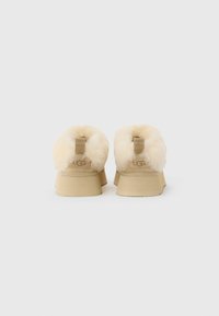 Zapatos slip-on UGG beige con suelas de goma gruesas y forro de piel de oveja crema esponjosa visible desde atrás sobre un fondo blanco.