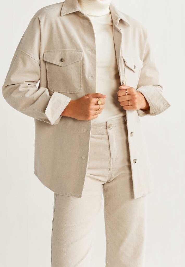 Personne portant une veste et un pantalon en velours côtelé beige avec un pull à col roulé blanc, tenant le devant de la veste avec les deux mains.