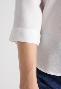 White, textured fabric blouse with rolled cuffs, showing part of a forearm. Blue denim jeans partially visible at the bottom.