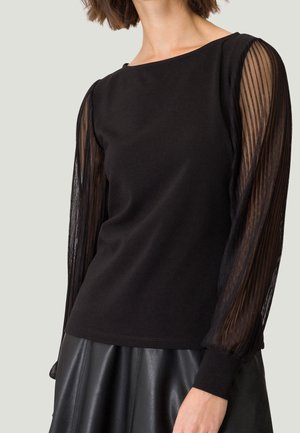 Kobieta w czarnej bluzce z przezroczystymi, plisowanymi długimi rękawami i czarnej skórzanej spódnicy na jednolitym tle.