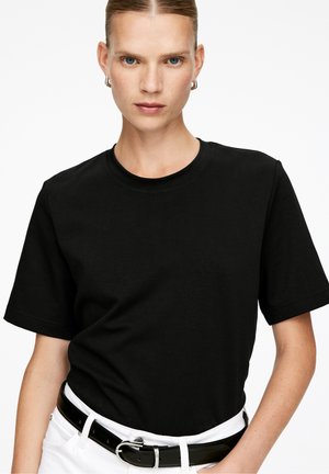 Czarna bawełniana koszulka z okrągłym dekoltem i krótkimi rękawami, w połączeniu z białym paskiem i widoczną klamrą paska, na jednolitym tle.