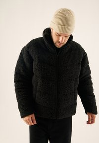 Schwarze Kunstpelzjacke mit hohem Kragen, Reißverschluss und strukturierter Oberfläche. Kombiniert mit einer beigen Strickmütze und schwarzen Hosen.