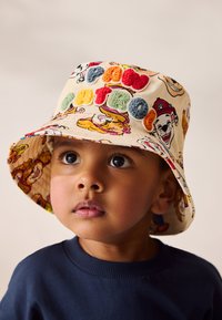 Eimerhut aus hellbeigem Stoff mit bunten Charakterdesigns und plüschigen Buchstaben. Breite Krempe und weiche Textur mit verspielten Mustern.