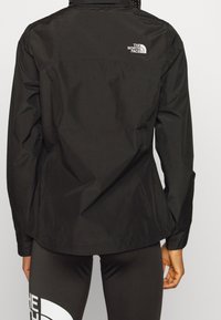 Persoon in een zwarte North Face jas en zwarte leggings met een groot wit North Face logo op de linkerdij, weergegeven van achteren.