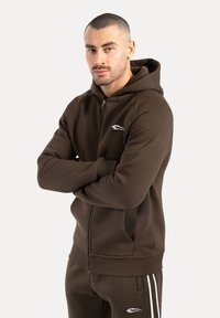 Sudadera con cremallera de color marrón, con capucha, puños acanalados y bolsillos frontales; presenta un pequeño logo blanco en el pecho; tejido suave y texturizado.