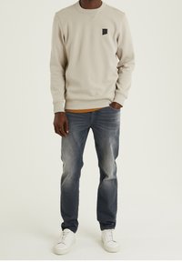 Světlé béžové mikina s kulatým výstřihem a malým černým logem, kombinovaná s vybledlými džínami a bílými teniskami.