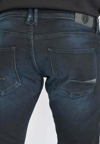 Vue de dos d'un jean en denim bleu foncé avec deux poches arrière et un petit patch de marque sur la ceinture.