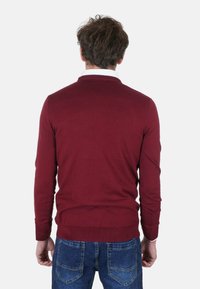 Maglione bordeaux con maglia strutturata, scollo rotondo e maniche lunghe. Abbinato a jeans di denim blu. Focus sul design posteriore e sulla vestibilità.