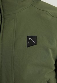 Khaki zelená bunda s hladkou texturou, obsahující černou nášivku s bílým logem. Obsahuje viditelné švy a zipové uzavření.