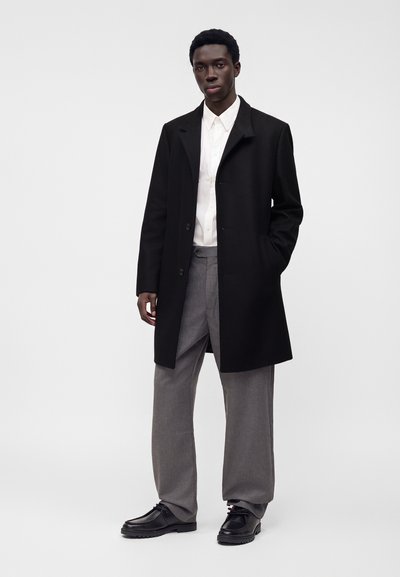 Homme portant un manteau noir, une chemise blanche boutonnée, un pantalon gris et des chaussures noires, debout avec une main dans la poche.