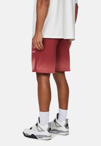 Pantaloni corti rossi con un design a gradiente, caratterizzati da elementi grafici bianchi. Abbinati a sneakers bianche e grigie, che mostrano dettagli testurizzati.