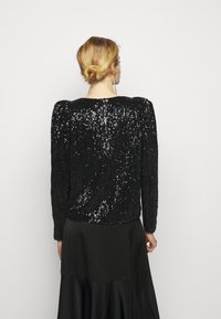 Kobieta z blond włosami związanymi w kok, ubrana w czarną koszulę z długim rękawem pokrytą cekinami i czarną satynową spódnicę, stoi przodem do gładkiej białej ściany.