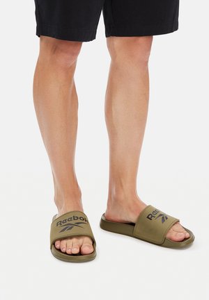 Nogi w czarnych szortach i oliwkowozielonych klapkach Reebok na białym tle.