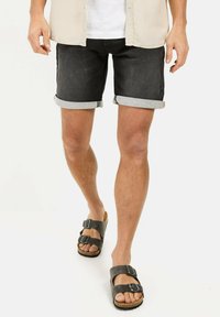 Schwarze Denim-Shorts mit umgeschlagenen Säumen, kombiniert mit grauen Sandalen mit doppelten Schnallen. Das Material wirkt robust mit einer leichten Textur.