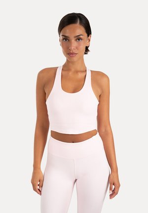 Donna con capelli scuri indossa un reggiseno sportivo rosa chiaro abbinato e leggings a vita alta, in piedi contro uno sfondo bianco semplice.