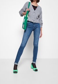 Cárdigan gris de punto con botones, camiseta negra, jeans skinny azules, sandalias de tacón verdes y calcetines negros con estampado; bolso verde con diseño floral.