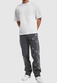 Hombre con una camiseta blanca sencilla, jeans gris oscuro con tres parches de cruces deshilachados, y zapatillas blancas, de pie con las manos detrás de la espalda.