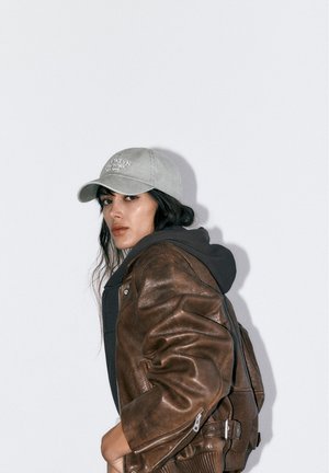 Frau mit grauer Mütze, brauner Lederjacke über einem schwarzen Kapuzenpullover, die über ihre Schulter blickt, vor einfachem hellgrauem Hintergrund.