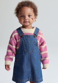 Denim-haal dress med brede skulderstropper og sideknapper, båret over en rosa og kremfarget stripet genser. Krøllete hår og nøytral bakgrunn.