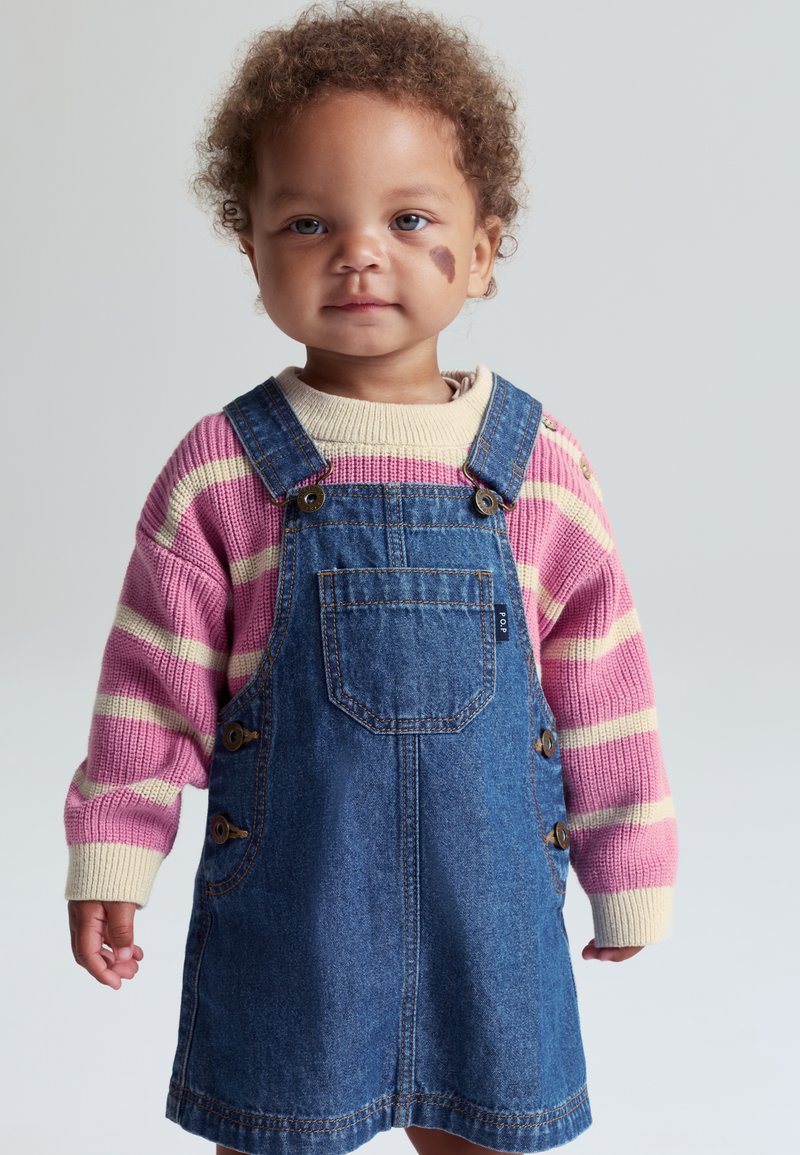 Denim-haal dress med brede skulderstropper og sideknapper, båret over en rosa og kremfarget stripet genser. Krøllete hår og nøytral bakgrunn.