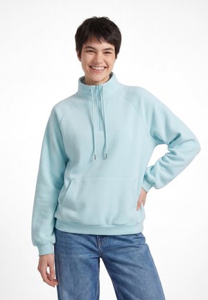 Giovane sorridente con capelli corti e scuri che indossa una felpa azzurro chiaro con zip e jeans blu, posa con una mano sui fianchi.
