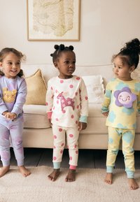 Trois enfants portent des pyjamas colorés : violet avec un motif de lion, crème avec des pois roses et une licorne, et jaune avec des accents de fleurs bleues.