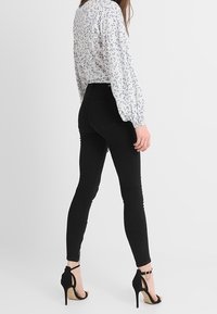 Jean skinny noir à taille haute, associé à un haut à motifs floraux. Modèle portant des talons aiguilles noirs, vu de dos.
