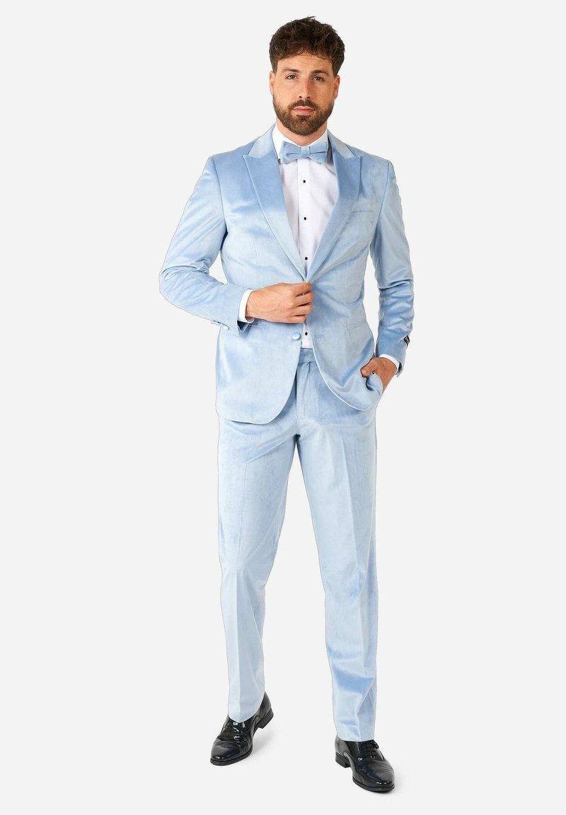 Traje de terciopelo azul claro con un corte ajustado, solapas con muesca y una camisa blanca con botones negros y una pajarita a juego. Zapatos de cuero negros.
