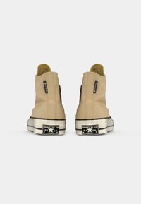 Baskets montantes beiges en toile avec une finition texturée ; elles présentent des accents noirs, une semelle en caoutchouc noire, et un logo Converse à l'arrière.