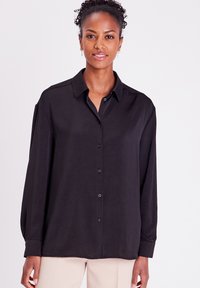 Chemise noire à manches longues avec col, confectionnée en tissu lisse ; présente des boutons sur le devant et une coupe décontractée.
