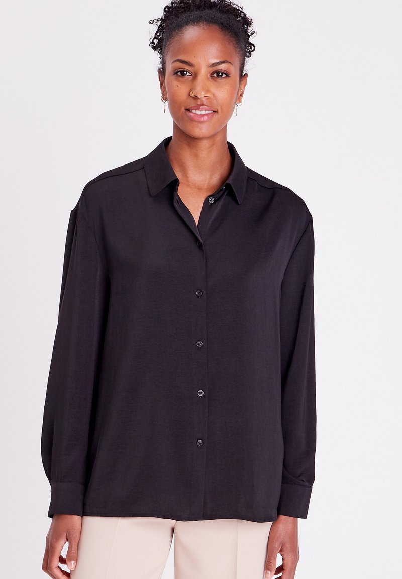 Chemise noire à manches longues avec col, confectionnée en tissu lisse ; présente des boutons sur le devant et une coupe décontractée.