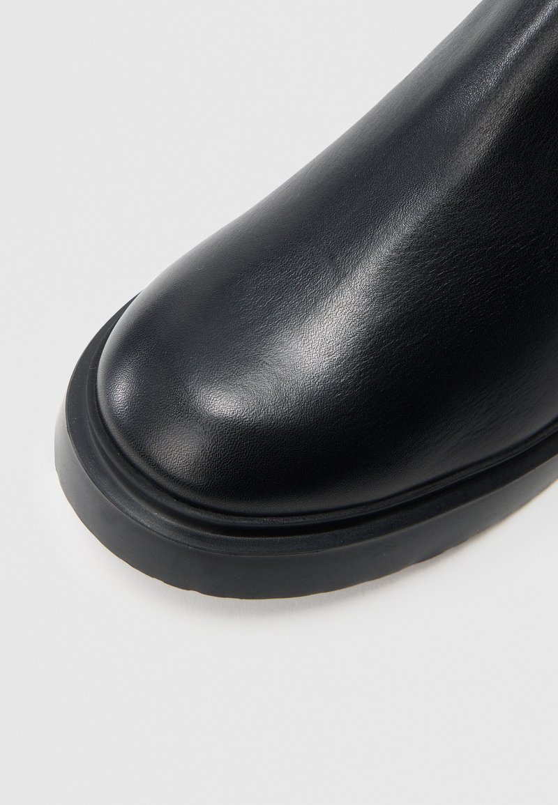 Svart ankelstövel i läder med rundad tå och texturerad yta, utrustad med en tjock svart gummisula och subtila sömmar.