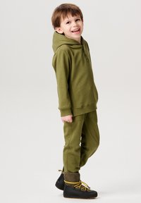 Olivgrüner Hoodie mit einer Knopfleiste an der Vorderseite und passenden Hosen. Strukturstoff mit gerippten Bündchen, kombiniert mit schwarzen Stiefeln mit gelben Schnürsenkeln.