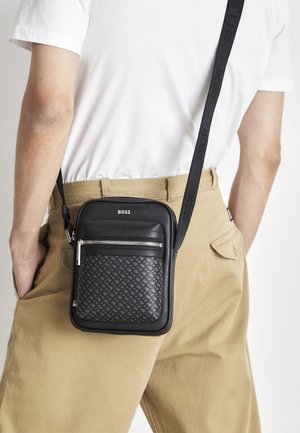 Persona che indossa pantaloni beige e camicia bianca con una borsa a tracolla nera con una tasca anteriore decorata e il logo "BOSS".
