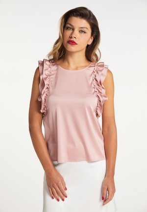 Frau trägt eine ärmellose hellrosa Bluse mit Rüschen an den Seiten und einen weißen Rock, steht vor einem schlichten weißen Hintergrund.