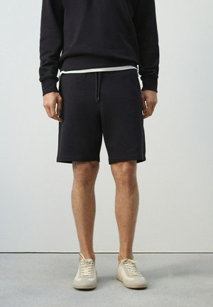 Hombre con pantalones cortos negros con cordón, camiseta negra de manga larga, camiseta interior blanca y zapatillas beige, de pie contra un fondo liso.