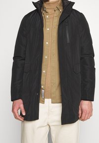 Veste imperméable noire avec capuche, fermeture éclair à l'avant, deux poches latérales et une poche poitrine zippée supplémentaire. Portée par-dessus une chemise beige.