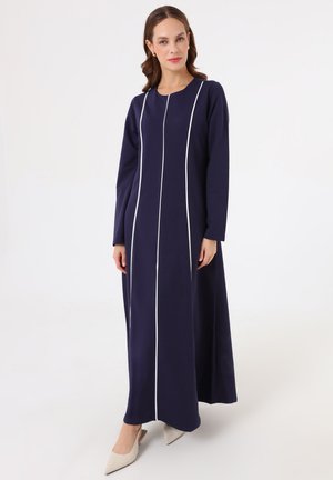Donna con capelli castani indossa un lungo vestito blu navy con strisce verticali bianche e tacchi beige appuntiti, in piedi davanti a uno sfondo bianco uniforme.