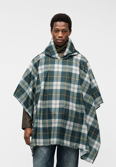 Homme portant un poncho à carreaux bleu, blanc et jaune avec une capuche par-dessus une veste marron et un jean bleu, debout devant un fond uni.