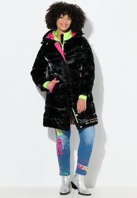 Cappotto puffo nero con finitura lucida, dettagli in rosa e verde neon, due tasche frontali, indossato sopra jeans con grafiche e stivaletti argento alla caviglia.