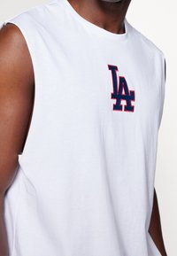 Bílý bezrukávový tričko vyrobené z bavlny, s vyšitým logem "LA" v červené a námořnické modré barvě na přední straně. Hladká textura s kulatým výstřihem.